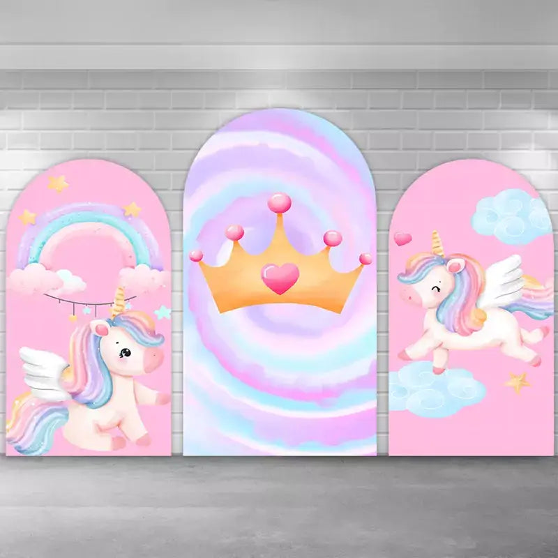 Kids’ unicorn party arch backdrop trio with soft pink backgrounds, winged unicorn illustrations, floating clouds, pastel rainbow arc, and a central cotton-candy swirl design topped with a golden crown and heart jewel.