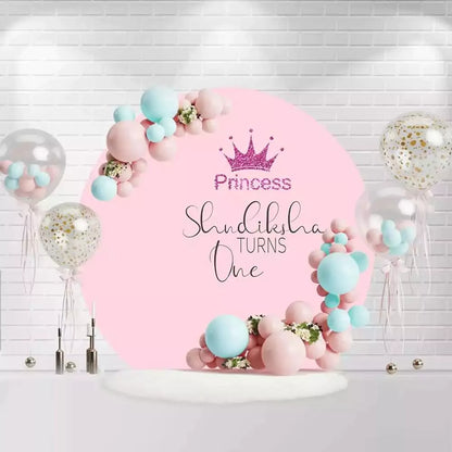 Round princess birthday backdrop with pink background, crown, and "Shundiksha Turns One" text
