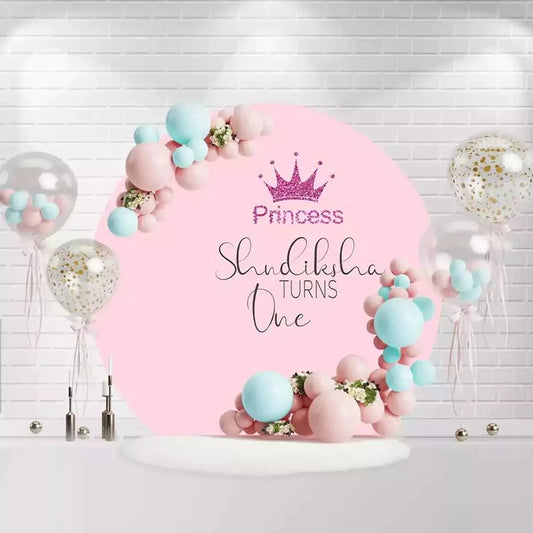 Round princess birthday backdrop with pink background, crown, and "Shundiksha Turns One" text
